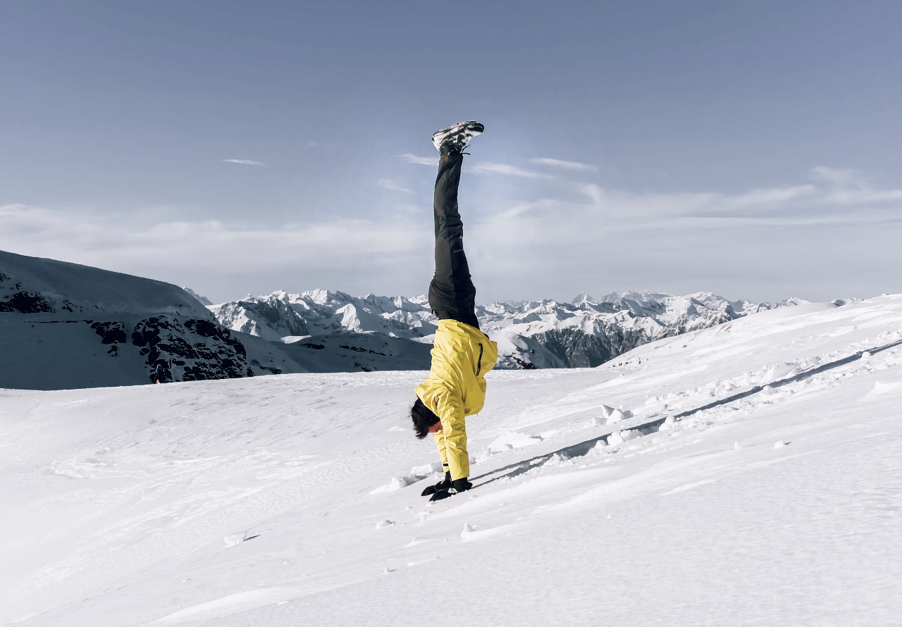 Persona in giacca gialla in equilibrio sulle mani (handstand) su un pendio innevato con panorama alpino