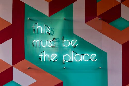 Tre Motivi per Fare Yoga – This Must Be the Place Insegna al neon this must be the place su sfondo turchese con geometrie decorative rosse e rosa