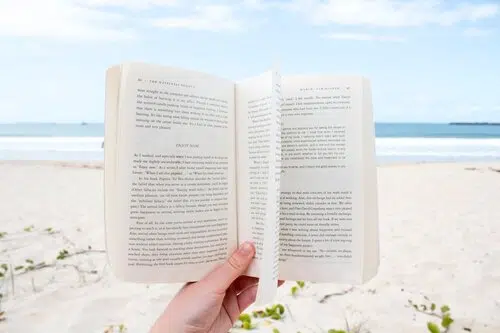 Mano che regge un libro aperto davanti a una spiaggia bianca con mare azzurro sullo sfondo