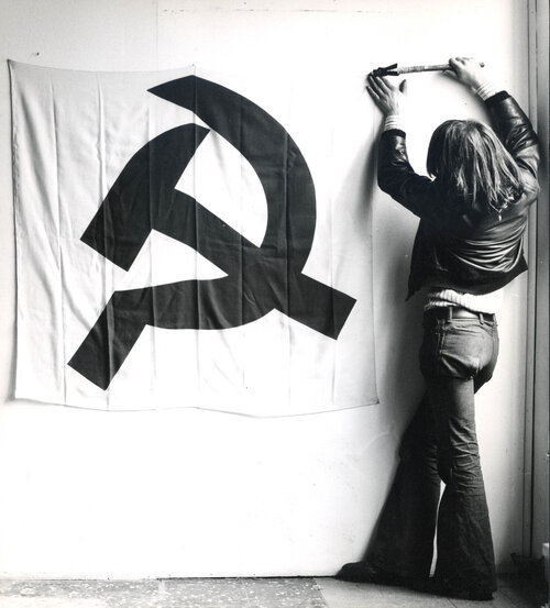 Fotografia in bianco e nero: persona vista di spalle appende uno striscione con falce e martello su parete urbana