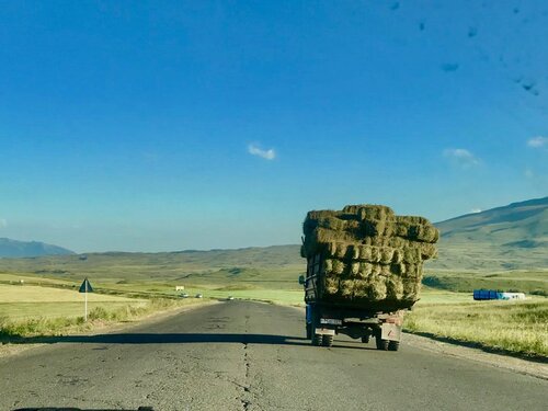 Camion Agricolo con Balle di Fieno su Strada Rurale Camion agricolo carico di balle di fieno su una lunga strada rurale dritta, montagne all'orizzonte