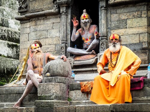 Tre sadhu con abiti arancioni e segni rituali sul corpo, seduti sui gradini di un tempio in pietra scolpita