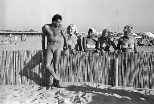 Fotografia vintage in bianco e nero: persona in costume da bagno appoggiata a recinzione di bamb&ugrave;, bagnanti sullo sfondo
