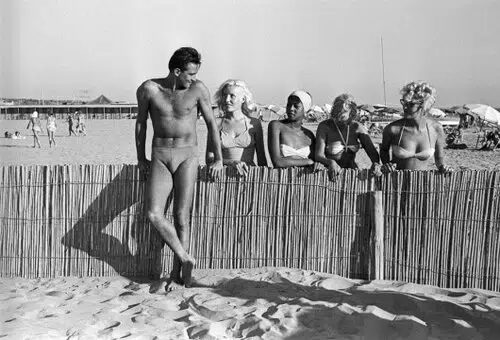 Fotografia vintage in bianco e nero: persona in costume da bagno appoggiata a recinzione di bamb&ugrave;, bagnanti sullo sfondo