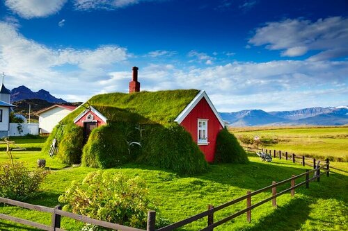 Casa tradizionale con tetto di torba verde e pareti rosse in un paesaggio nordico, montagne e cielo azzurro sullo sfondo