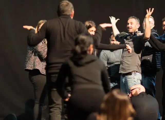 Gruppo di persone in un esercizio di improvvisazione teatrale, braccia aperte e gesti espressivi su sfondo scuro