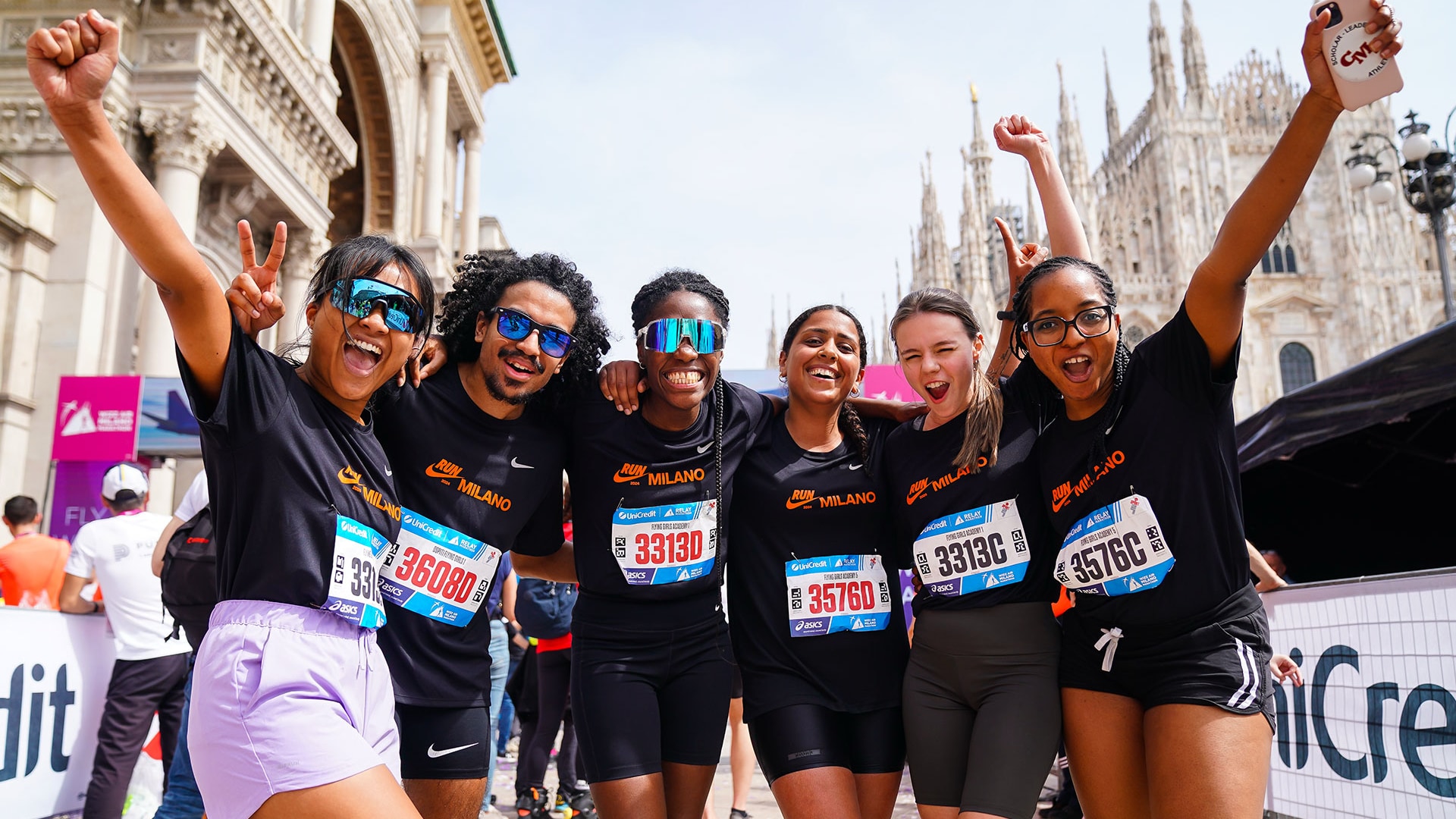 Sei corridori in magliette nere 'RUN MILANO' esultano con le braccia alzate davanti al Duomo di Milano
