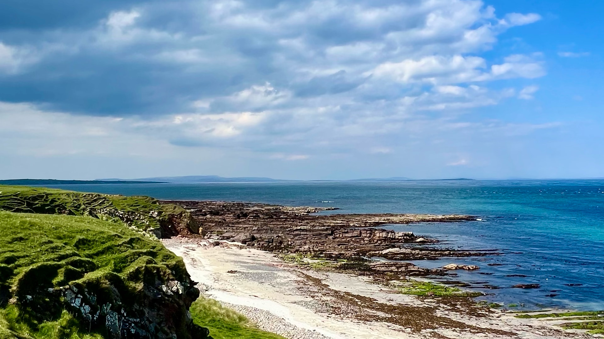John O’Groats – Paesaggio costiero della Scozia settentrionale Costa rocciosa con erba verde, ciottoli bianchi e mare azzurro sotto un cielo nuvoloso, paesaggio costiero nordico