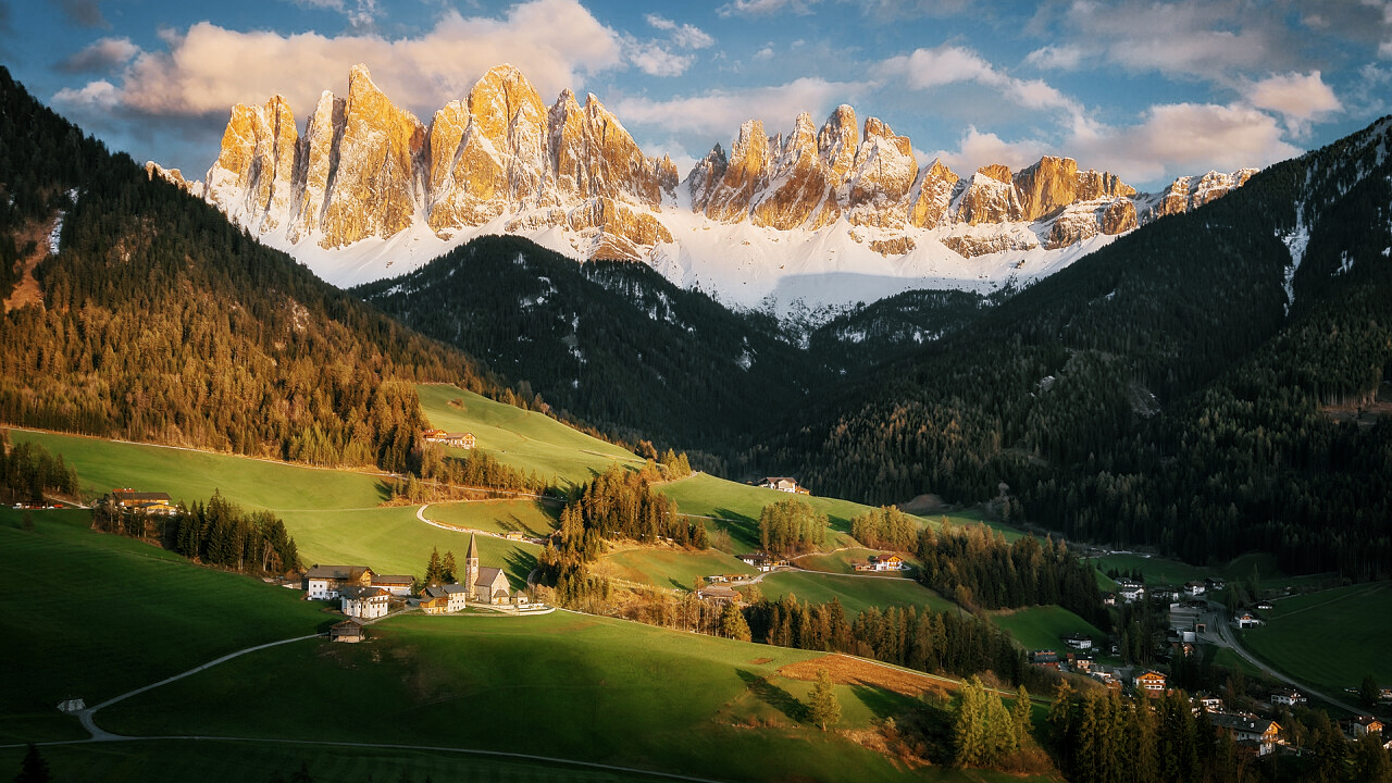 Panorama dolomitico con vette frastagliate illuminate dalla luce calda, valle verde e piccolo borgo con chiesa