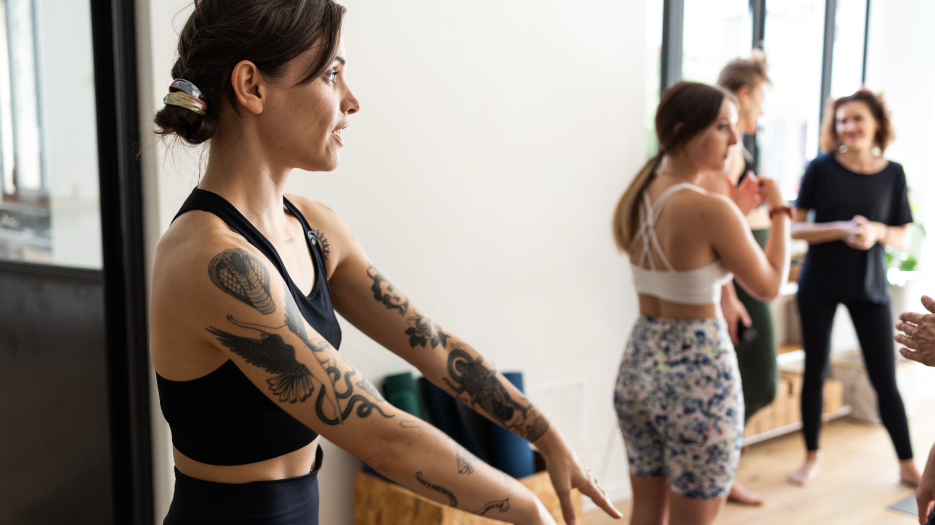 Lezione di Yoga – Insegnante con Tatuaggi Guida la Classe Insegnante con tatuaggi sulle braccia guida una lezione di yoga in studio luminoso, allievi sullo sfondo