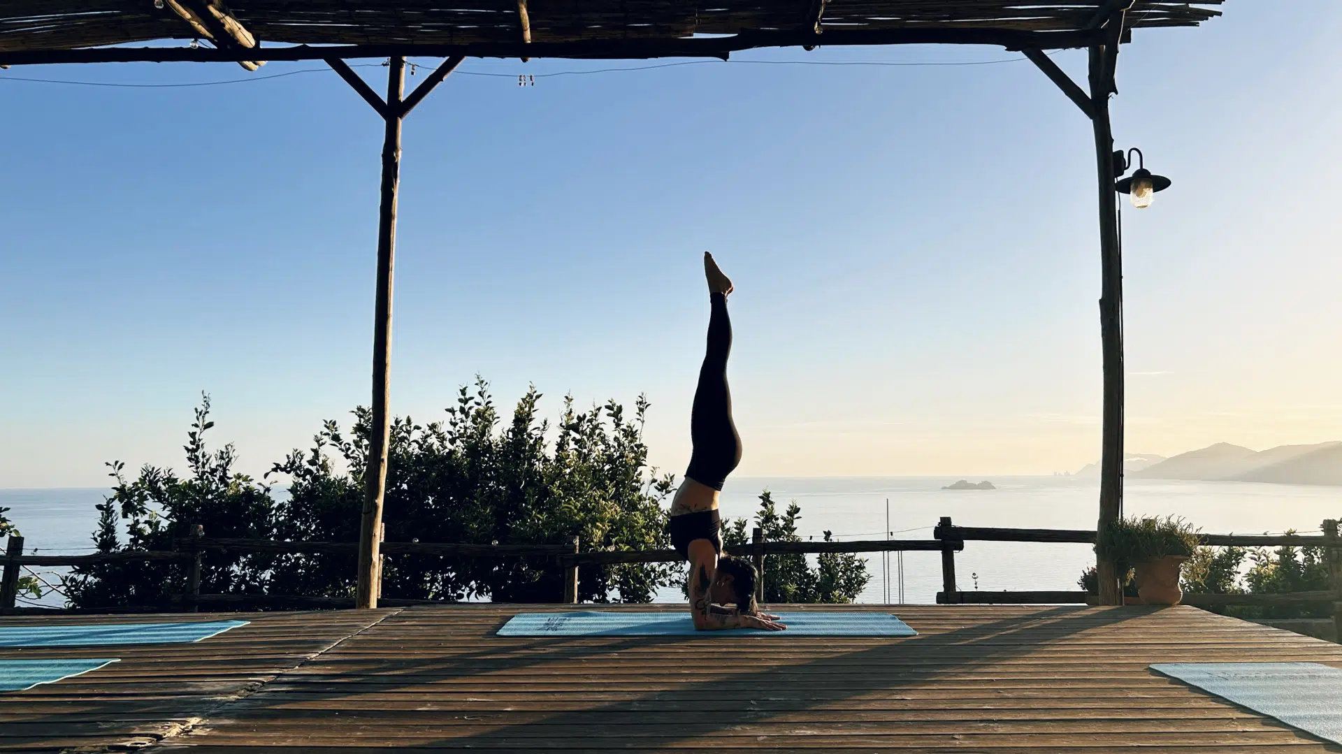 Insegnante di yoga in equilibrio invertito su tappetino blu, su una terrazza in legno con vista sul mare